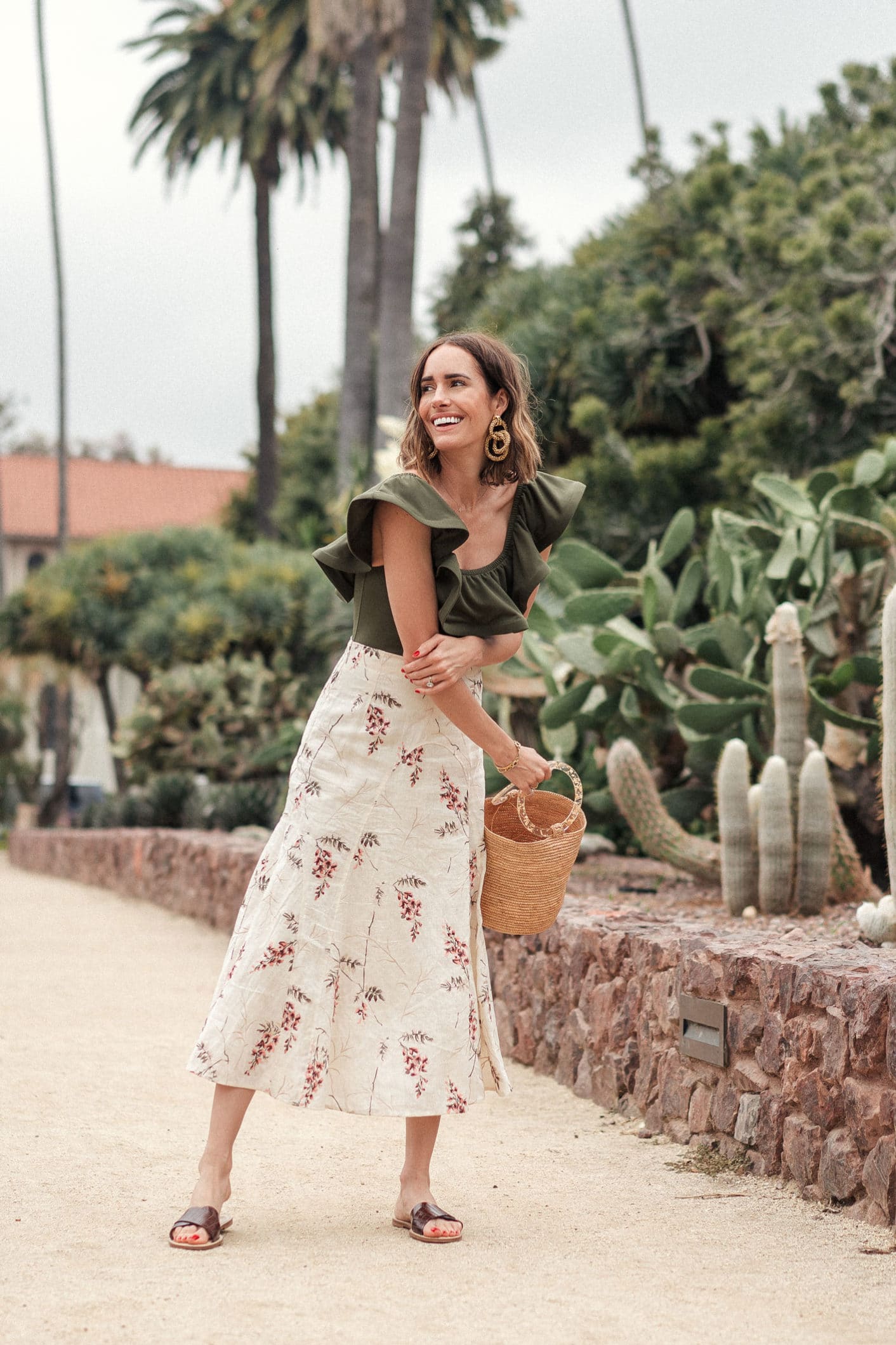 Woman in a floral summer midi skirt and olive top, smiling with a basket, surrounded by greenery in a sunny setting.